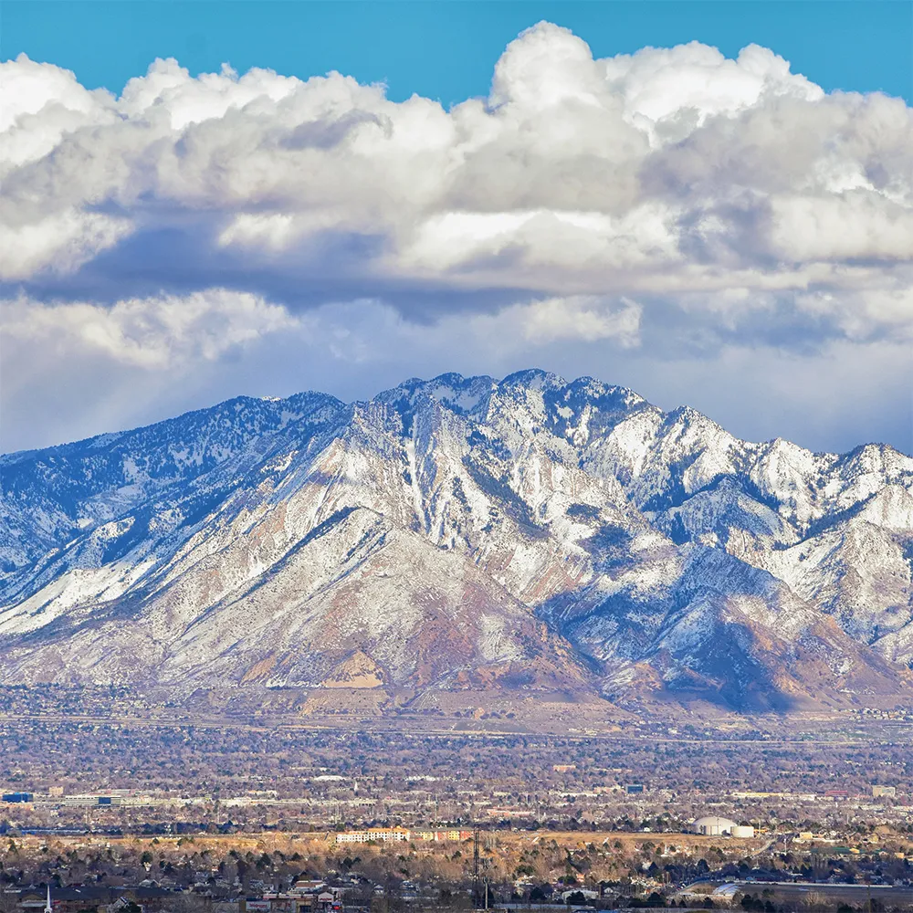 Snow-capped Wasatch Mountains rising above an urban valley with dramatic white clouds in a blue sky.